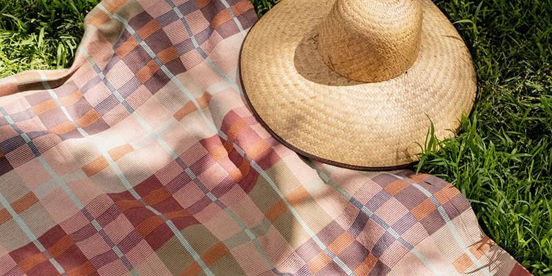 A Mungo picnic blanket in a pink and rust check spread on sunlit grass with a straw sun hat