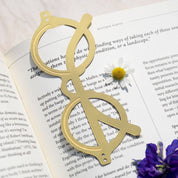 OCTAEVO The Voyage brass bookmark resting on an open book beside a daisy