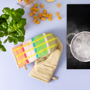 Checked oven mitts on a kitchen counter beside fresh basil and a pot of boiling water