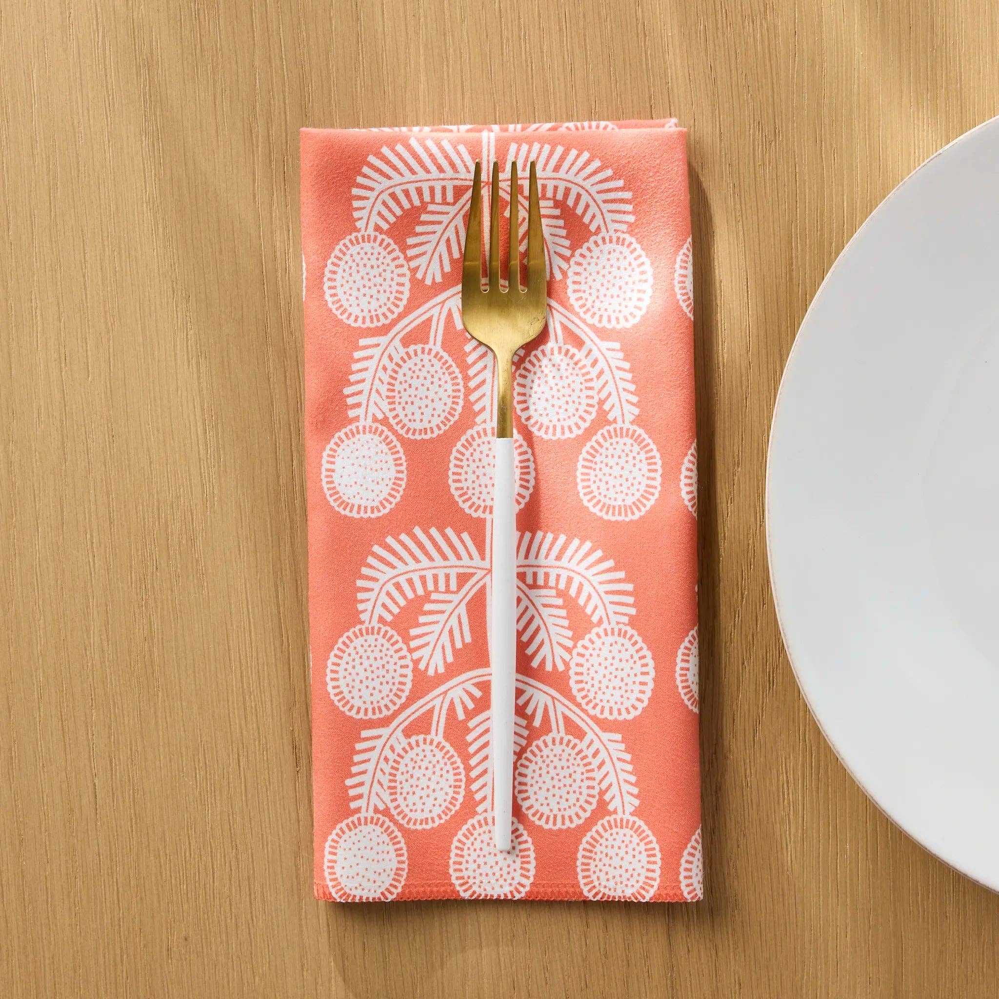Graphic Daisies dinner napkin folded on a wooden table with a gold fork, white daisy sprigs on coral