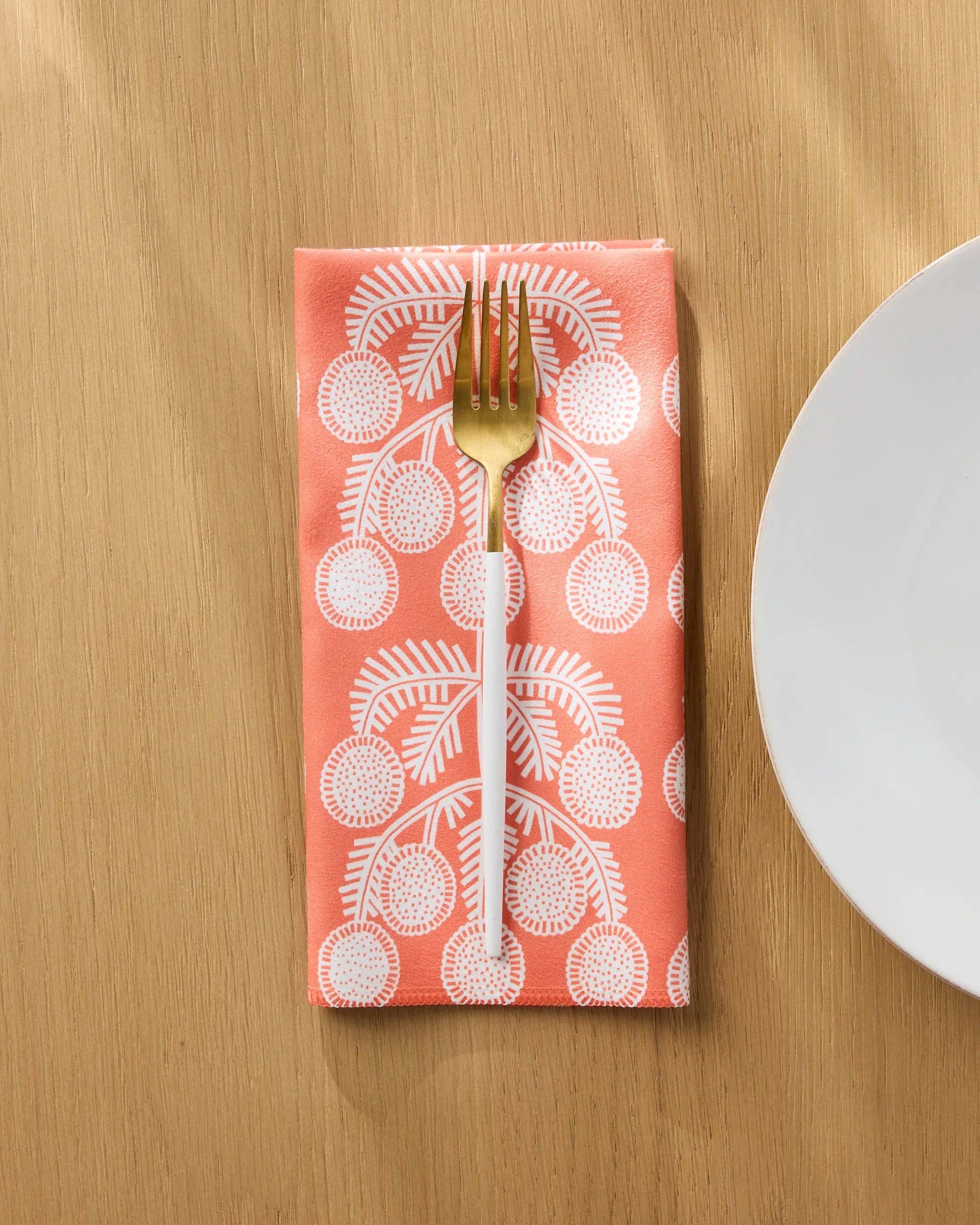 Graphic Daisies dinner napkin folded on a wooden table with a gold fork, white daisy sprigs on coral