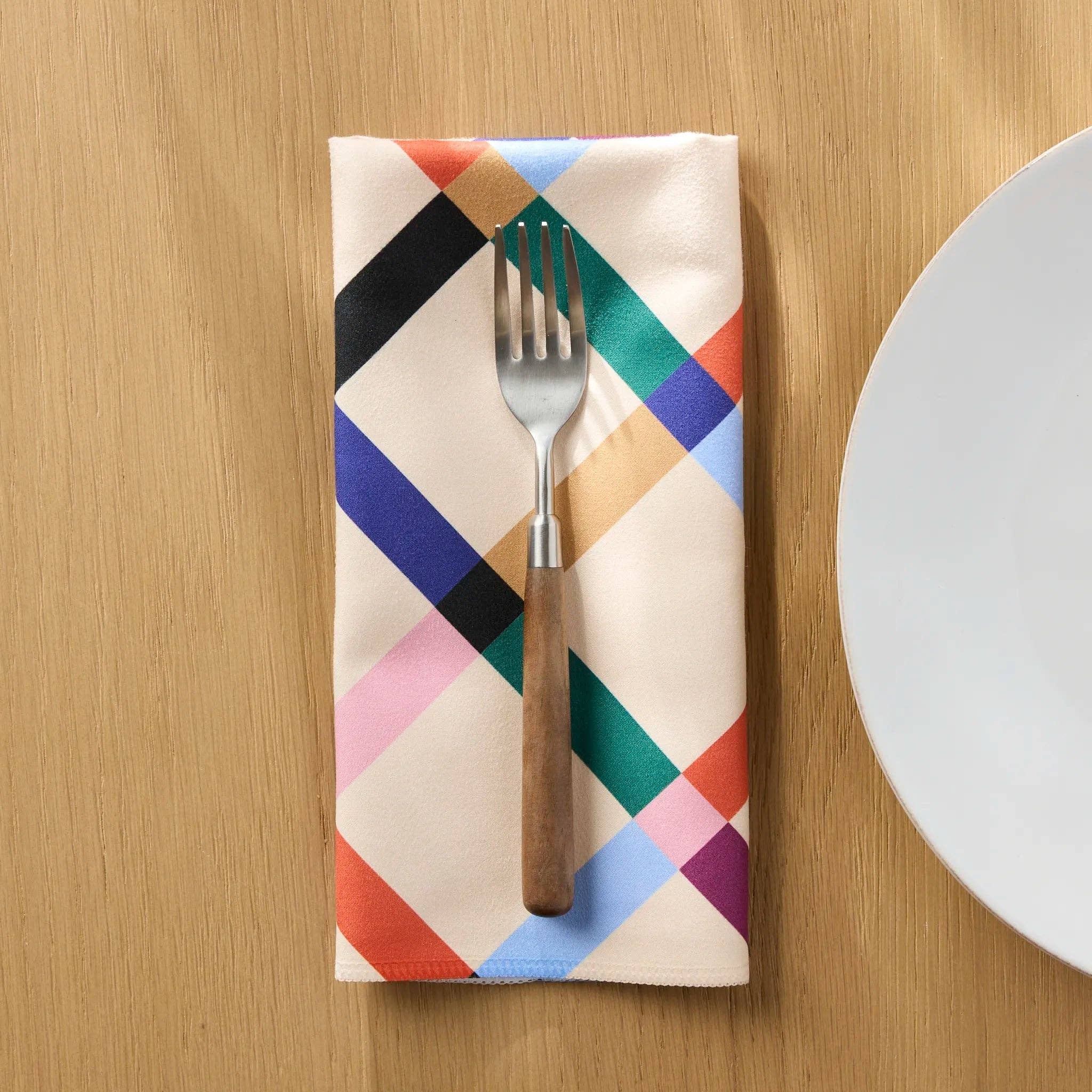 Cool Grid dinner napkin folded on a wooden table beside a plate and fork, diagonal lattice in black, green, blue and coral on cream