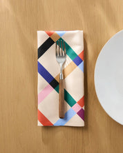 Cool Grid dinner napkin folded on a wooden table beside a plate and fork, diagonal lattice in black, green, blue and coral on cream