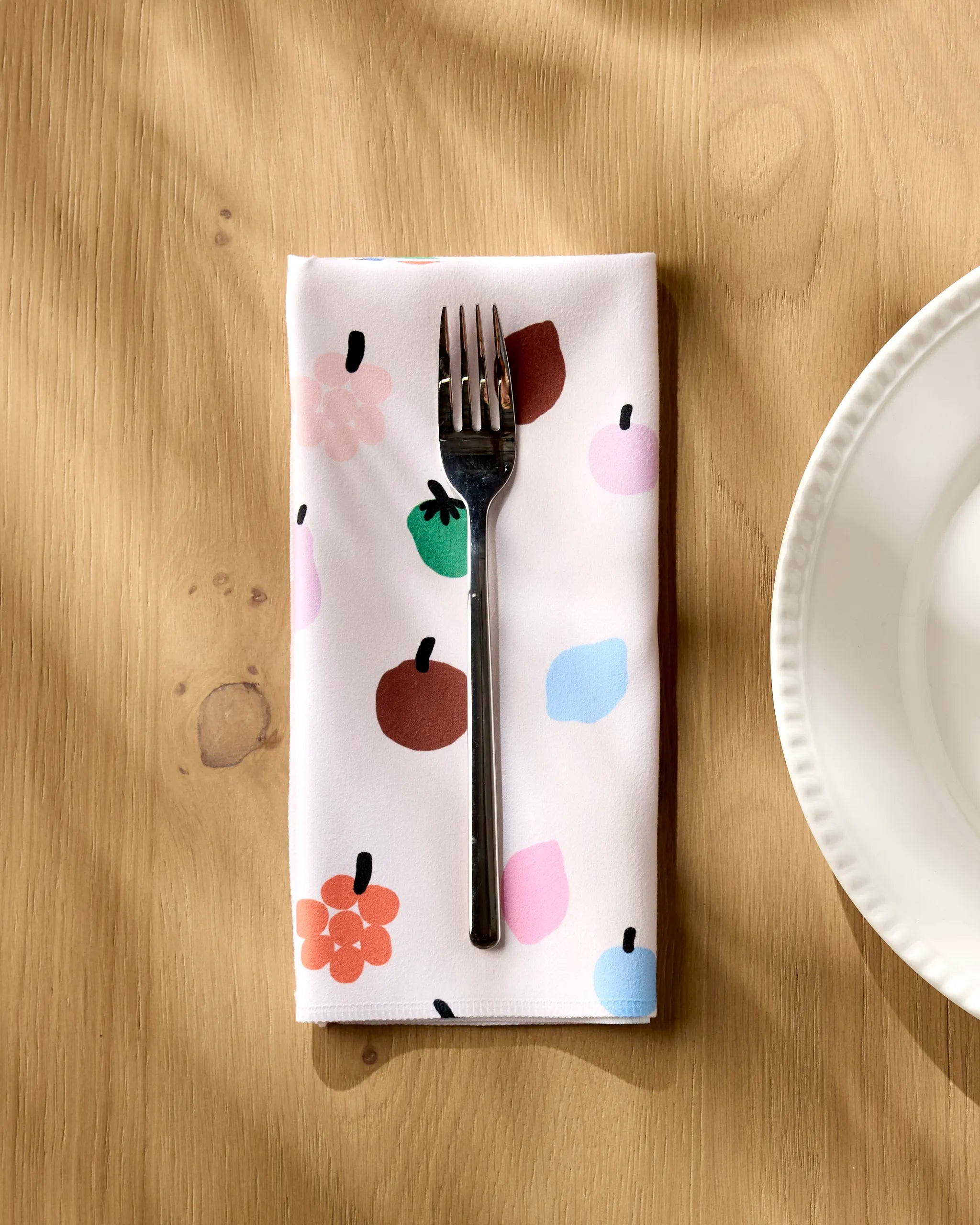 Fruit Basket napkin folded on a wooden table with a fork resting on top, beside a white plate