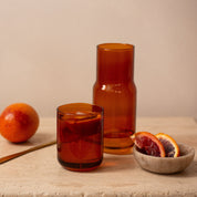 Amber tinted carafe and matching glass filled with blood orange juice, beside sliced citrus
