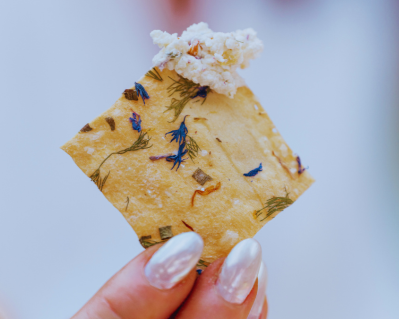 A hand holding a single No. 1 cracker topped with cheese, edible flowers visible through the crisp