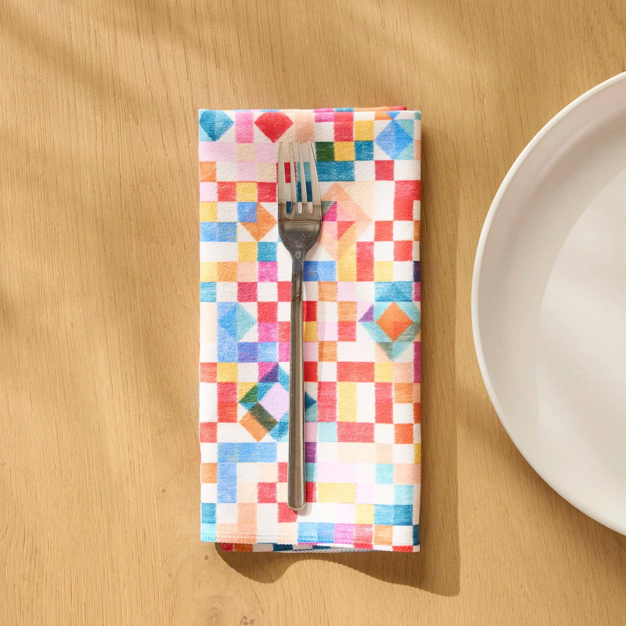 Patchwork Geo dinner napkin folded on a wooden table beside a plate and fork, watercolour squares in red, blue and green