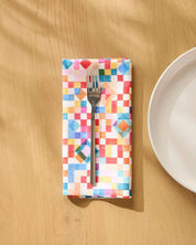 Patchwork Geo dinner napkin folded on a wooden table beside a plate and fork, watercolour squares in red, blue and green
