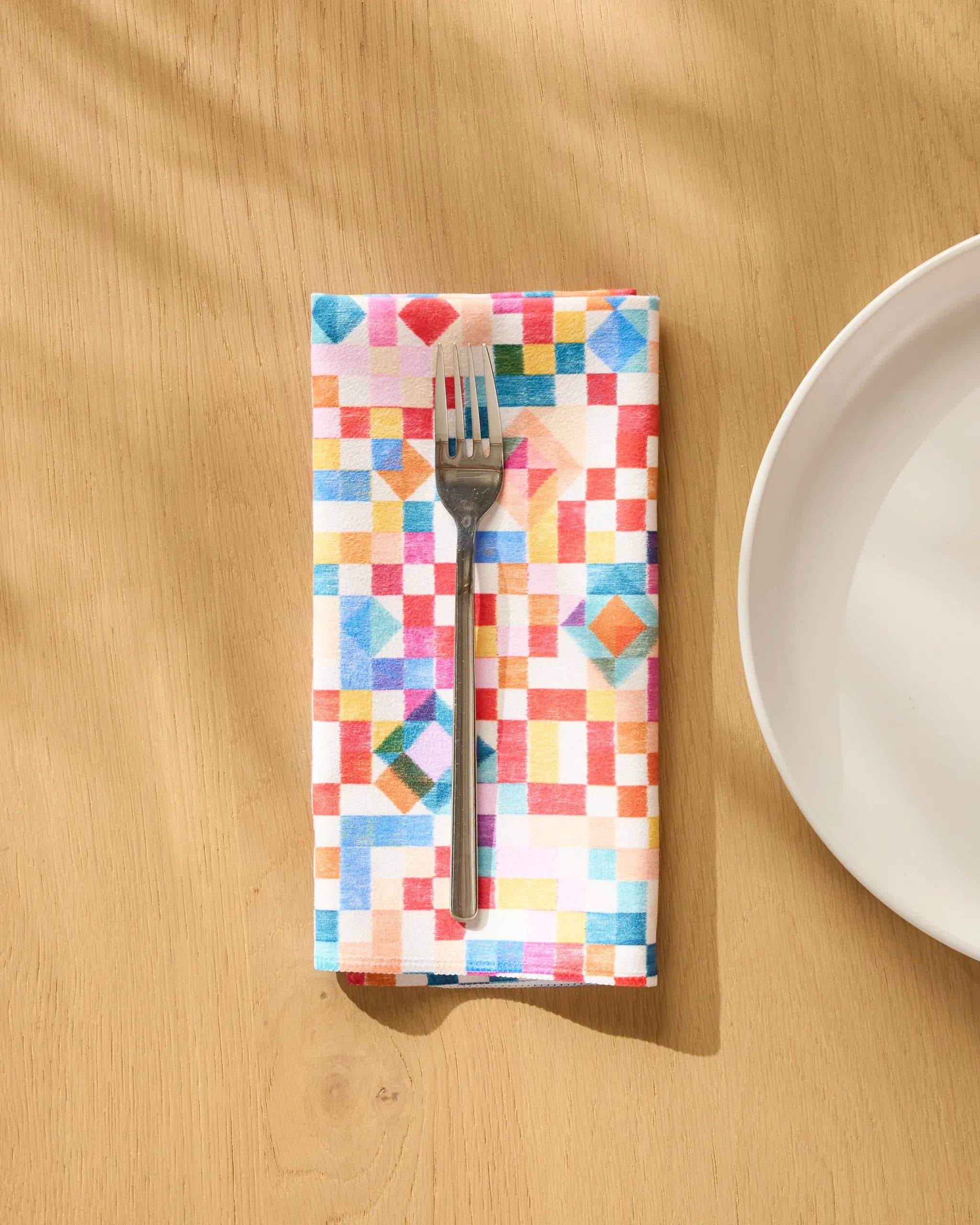 Patchwork Geo dinner napkin folded on a wooden table beside a plate and fork, watercolour squares in red, blue and green