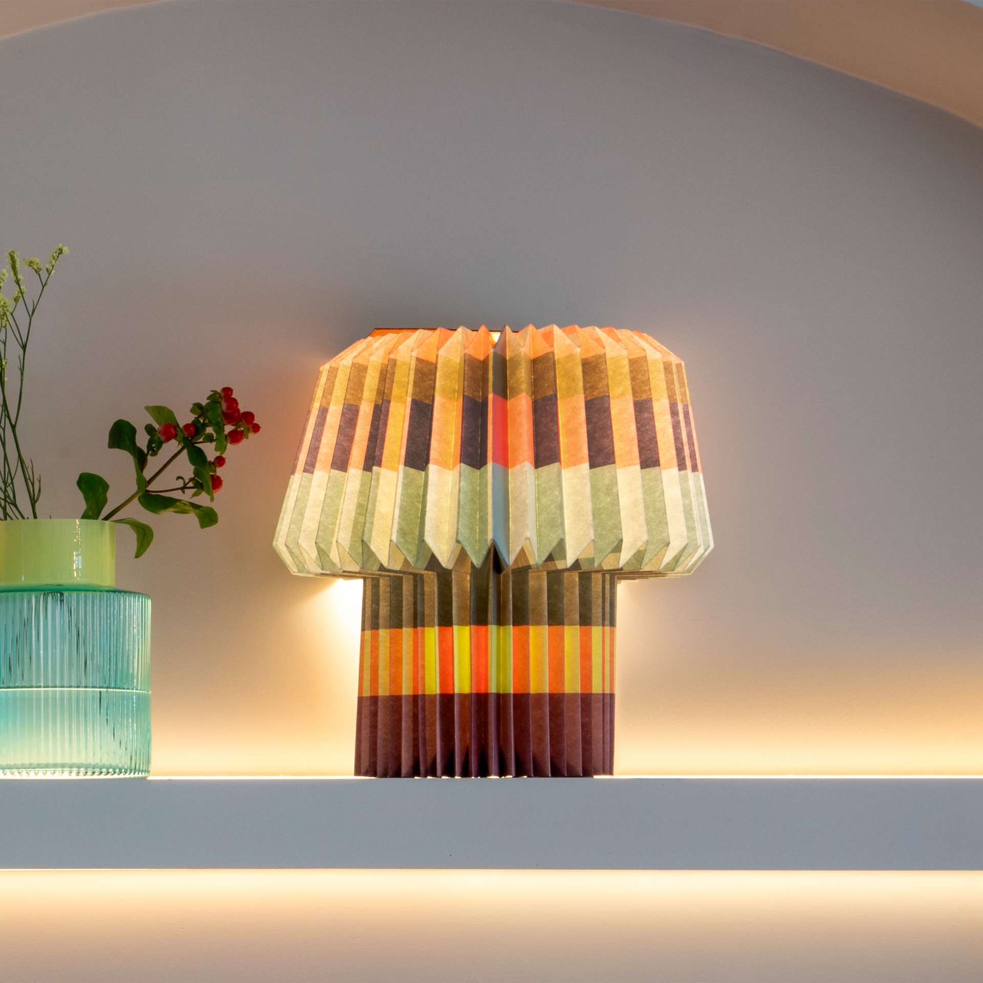 The blocks lamp lit on a pale shelf next to a green vase of berried stems