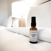 Los Poblanos Lavender Mist in an amber glass bottle with a black pump, on white bedding
